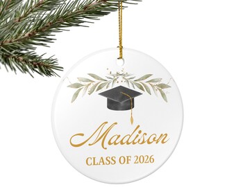 May include: White ceramic graduation ornament with a black mortarboard, gold tassel, and the name "Madison" in gold script. The ornament is decorated with green and gold foliage and the text "CLASS OF 2026". A gold string is attached for hanging.