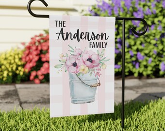 May include: A personalized garden flag with the text "THE Anderson FAMILY" above a watercolor illustration of pink flowers in a blue bucket. The flag has a pink and white striped background and is displayed on a black metal stand.
