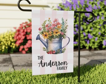 May include: A garden flag featuring a watercolor illustration of a blue watering can overflowing with colorful flowers. The flag has a pink and white checkered background and the text "The Anderson Family". The flag is displayed on a black metal stand.