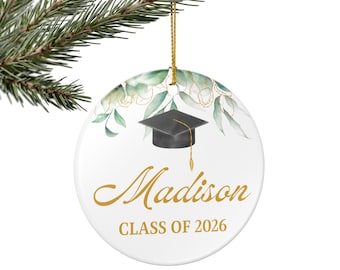 May include: A white ceramic graduation ornament with a gold string for hanging. The ornament features a black graduation cap, the name "Madison" in gold script, and "CLASS OF 2026" in gold. Green and gold floral accents decorate the top.