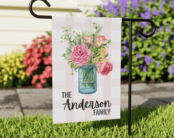 May include: A garden flag with a pink and white striped background. The flag features a watercolor illustration of a bouquet of pink and yellow roses in a blue jar. The text "The Anderson Family" is below the illustration.