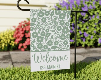 Könnte beinhalten: Eine Gartenflagge mit einem floralen Design in Salbeigrün und Weiß. Die Flagge trägt die Aufschrift "Welcome" und "123 MAIN ST" in einer Schriftart. Die Flagge wird auf einem schwarzen Metallständer in einem grasbewachsenen Garten gezeigt.