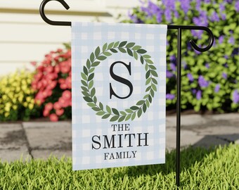 Könnte beinhalten: Eine Gartenflagge mit hellblauem und weißem Karomuster. Sie zeigt den Text "THE SMITH FAMILY" unter einem grünen Kranz, der einen schwarzen Buchstaben "S" umschließt. Die Flagge ist auf einem schwarzen Metallständer in einem Garten befestigt.