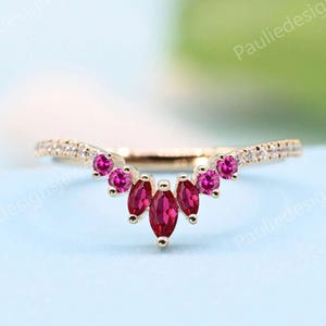 May include: A gold-toned ring with a curved design, featuring a cluster of marquise-cut, deep red gemstones at the center. Smaller, round red gemstones and tiny clear stones adorn the band, creating a sparkling effect. The ring is set against a light blue background.