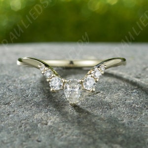 Puede incluir: Un delicado anillo de oro con un diamante de forma pera en el centro y diamantes redondos más pequeños a cada lado. El anillo está curvado en forma de V.