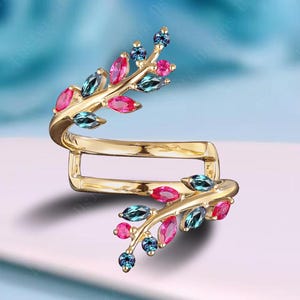 May include: A gold-colored ring with a leaf design. The ring features a series of colorful gemstones in shades of pink, blue, and teal. The gemstones are arranged to mimic leaves along the ring's design. The ring has a unique, open design.