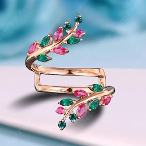 Op de afbeelding: Een roségouden ring met een bladvorm. De ring heeft groene en roze edelstenen die zijn gerangschikt om op bladeren te lijken. De ring wikkelt zich om de vinger met een uniek, open ontwerp. De edelstenen zijn smaragd en robijn van kleur.