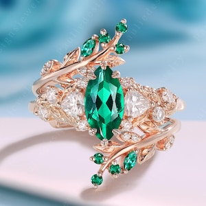 May include: A rose gold ring set featuring a marquise-cut emerald green gemstone as the centerpiece, surrounded by smaller white gemstones and green accents. The ring has a leaf-like design.