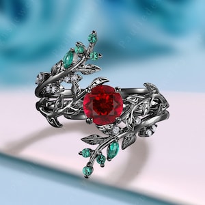 May include: Black ring featuring a red gemstone surrounded by smaller white gemstones. The ring has a design of intertwined branches with green leaves.