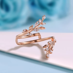 May include: A rose gold ring featuring a delicate design of leaves and small clear stones. The ring is open on one side and has a double band.