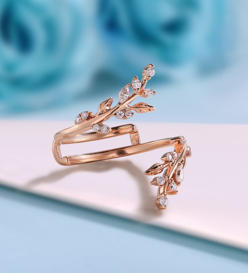 May include: A rose gold ring featuring a delicate design of leaves and small clear stones. The ring is open on one side and has a double band.