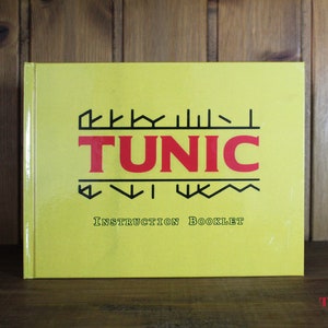 Puede incluir: Un folleto de instrucciones amarillo para el videojuego "TUNIC". El folleto tiene texto negro y rojo y un logotipo estilizado con el t&iacute;tulo del juego.