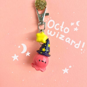 Può includere: Un ciondolo a forma di polpo rosa con un cappello da mago con stelle gialle e una chiusura color oro. Il ciondolo ha il testo "Octo Wizard!" su uno sfondo rosa con stelle e lune bianche.