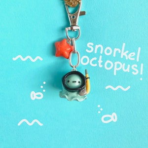 Könnte beinhalten: Ein türkisfarbener Oktopus mit einer schwarzen Tauchermaske und einem gelben Schnorchel. Der Oktopus ist an einem silbernen Schlüsselanhänger mit einem goldenen Blatt-Anhänger und einem roten Stern-Anhänger befestigt. Der Text "snorkel octopus!" ist in Weiß auf blauem Hintergrund geschrieben.