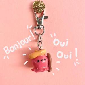 Pu&ograve; includere: Un portachiavi a forma di polpo rosa e giallo con una baguette marrone e un ciondolo a foglia d'oro. Il portachiavi ha le parole "Bonjour!" e "Oui Oui!" scritte intorno ad esso.