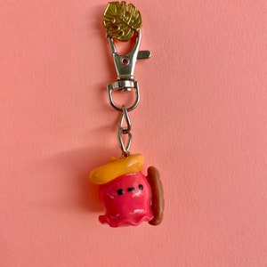 Pu&ograve; includere: Un portachiavi a forma di polpo rosa e giallo con un hot dog marrone e un ciondolo a foglia d'oro. Il polpo indossa un cappello giallo.