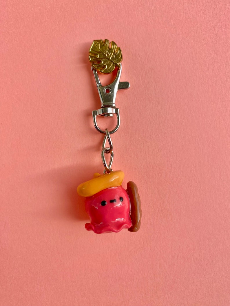 Pu&ograve; includere: Un portachiavi a forma di polpo rosa e giallo con un hot dog marrone e un ciondolo a foglia d'oro. Il polpo indossa un cappello giallo.