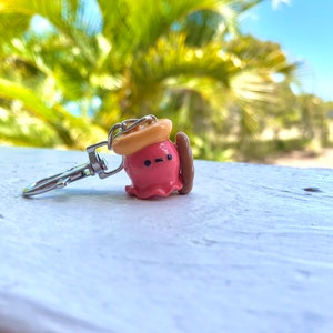 Pu&ograve; includere: Un portachiavi a forma di polpo rosa con un cappello giallo e una tavola da surf marrone. Il polpo ha una piccola bocca e occhi neri.