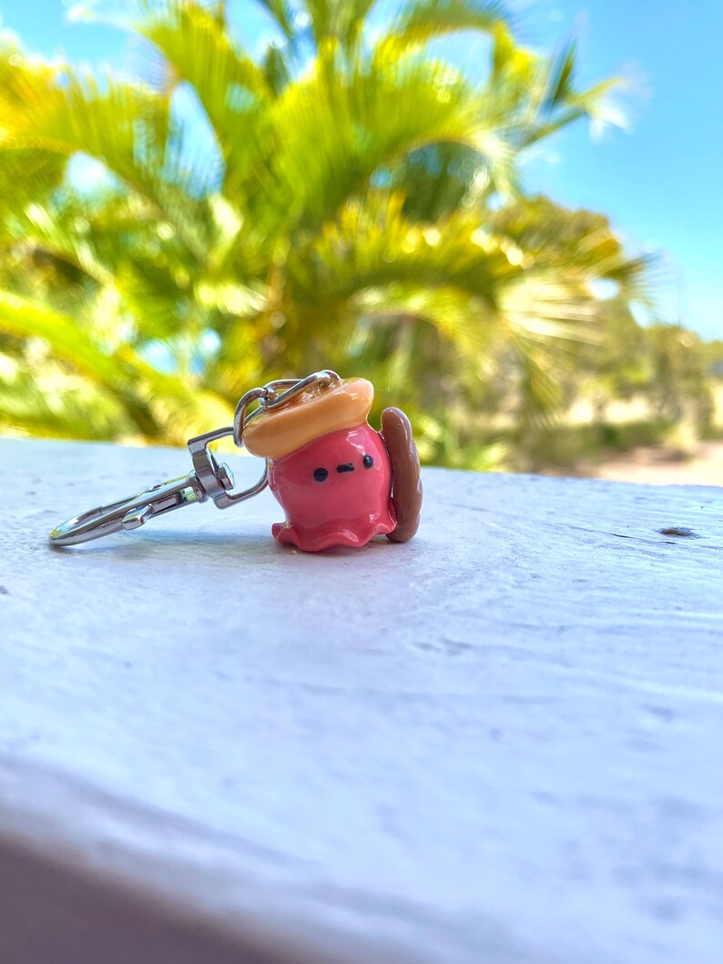 Pu&ograve; includere: Un portachiavi a forma di polpo rosa con un cappello giallo e una tavola da surf marrone. Il polpo ha una piccola bocca e occhi neri.