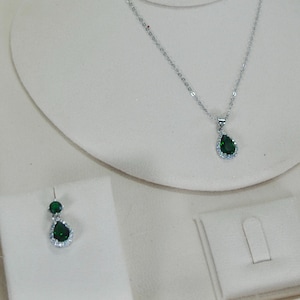 May include: A silver necklace with a teardrop-shaped green pendant, displayed with matching earrings. The pendant and earrings are surrounded by small, clear stones. The jewelry is presented on a white display.