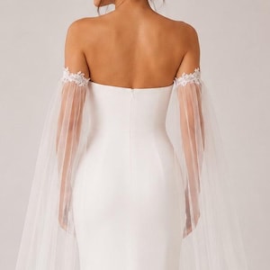 May include: A white, strapless wedding gown with a mermaid silhouette. The dress features sheer, flowing sleeves and delicate floral embellishments at the shoulders. The back of the dress is visible, showcasing the elegant design and the long train.