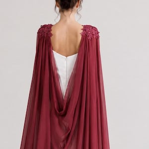 May include: A burgundy chiffon cape with floral lace shoulder accents. The cape drapes over a white dress, creating a flowing silhouette. The back of the dress is visible, with a V-shaped neckline. The overall style is elegant and suitable for special occasions.