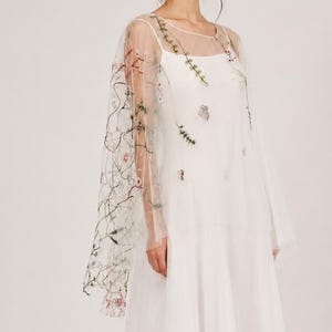 Könnte beinhalten: Ein weißes Brautkleid mit einem bestickten Überrock. Der Überrock ist mit zarten Blumenstickereien in Grün-, Rosa- und Brauntönen verziert. Das Kleid hat ein schlichtes, elegantes Design, für eine Braut.