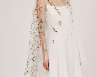 May include: A white wedding dress with a sheer overlay. The overlay is adorned with delicate floral embroidery in shades of green, pink, and brown. The dress has a simple, elegant design, perfect for a bride.