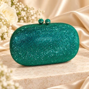 May include: An oval-shaped clutch bag completely covered in small, shimmering emerald green crystals. The bag features a silver-toned metal frame and a top closure mechanism consisting of two round, crystal-encrusted knobs that snap together.