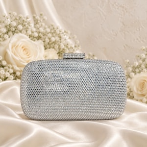 Puede incluir: Un bolso de mano rectangular plateado completamente cubierto de peque&ntilde;os cristales reflectantes. El bolso presenta un dise&ntilde;o de bordes redondeados y un cierre met&aacute;lico rectangular en la parte superior, tambi&eacute;n incrustado con una fila de cristales claros.