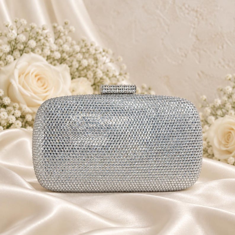 Puede incluir: Un bolso de mano rectangular plateado completamente cubierto de peque&ntilde;os cristales reflectantes. El bolso presenta un dise&ntilde;o de bordes redondeados y un cierre met&aacute;lico rectangular en la parte superior, tambi&eacute;n incrustado con una fila de cristales claros.