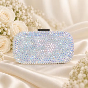 Puede incluir: Un bolso de mano rectangular completamente cubierto de cristales iridiscentes que reflejan la luz y brillan con tonos blancos y arco&iacute;ris. El bolso presenta un cierre rectangular negro s&oacute;lido en la parte superior central, creando un contraste marcado.