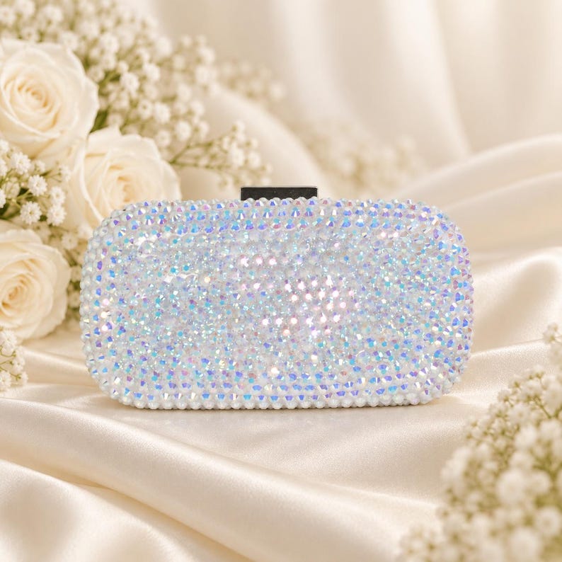 Puede incluir: Un bolso de mano rectangular completamente cubierto de cristales iridiscentes que reflejan la luz y brillan con tonos blancos y arco&iacute;ris. El bolso presenta un cierre rectangular negro s&oacute;lido en la parte superior central, creando un contraste marcado.