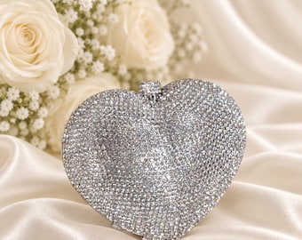 Puede incluir: Un bolso de mano en forma de corazón completamente cubierto de pequeños cristales plateados reflectantes. El accesorio cuenta con un marco de metal plateado y un pequeño cierre circular en la parte superior central, creando una apariencia muy texturizada y brillante.