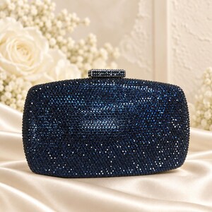 Puede incluir: Un bolso de mano rectangular completamente cubierto de cristales azul oscuro. La superficie presenta una textura densa y brillante en todo su conjunto. Un cierre rectangular en la parte superior tambi&eacute;n est&aacute; incrustado con cristales azul oscuro a juego, proporcionando un cierre seguro para este accesorio de noche estructurado.