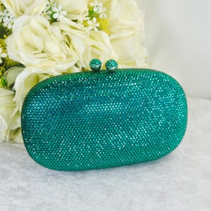 Peut inclure: Une pochette de forme ovale entièrement recouverte de petits cristaux vert émeraude scintillants. Le sac présente une structure rigide avec une fermeture à cliquet sécurisée sur le dessus, composée de deux boutons métalliques ronds incrustés.