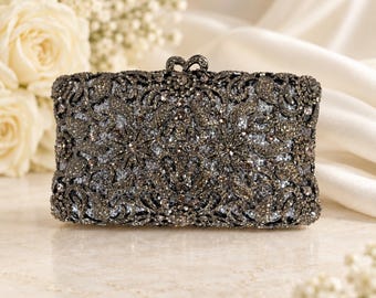 Puede incluir: Un bolso de mano rectangular con acabado en color plomo oscuro y un intrincado patrón de filigrana calada. La superficie está densamente incrustada con pequeños cristales y cuentas oscuras brillantes, con un cierre metálico decorativo.