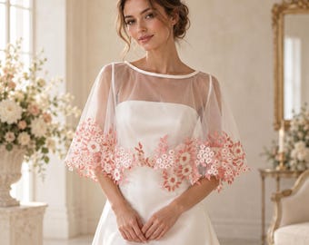 May include: A sheer white tulle bridal capelet featuring a delicate scalloped hem adorned with intricate pink floral embroidery. The garment has a wide boat neckline and short, flowing sleeves that drape over the shoulders of a satin dress.