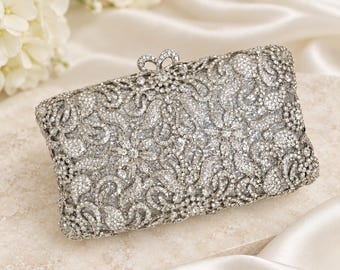 Puede incluir: Un bolso de mano rectangular plateado con un revestimiento de filigrana completamente incrustado de cristales transparentes brillantes. El bolso tiene un marco metálico y un pequeño cierre decorativo en el centro con un denso patrón floral.