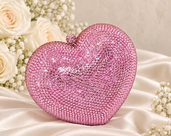 Puede incluir: Un bolso de mano en forma de corazón completamente cubierto de pequeños cristales rosas brillantes. El bolso cuenta con un pequeño cierre metálico incrustado con cristales en la parte superior central, diseñado para cerrar el accesorio rígido.