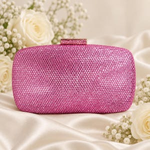 Puede incluir: Un bolso de mano rectangular completamente cubierto de peque&ntilde;os cristales de color rosa vibrante. El bolso cuenta con un cierre rectangular a juego, tambi&eacute;n incrustado con cristales rosas en la parte superior, para un aspecto uniforme.
