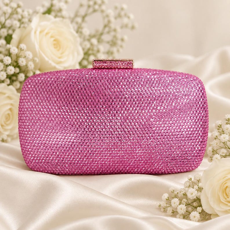 Puede incluir: Un bolso de mano rectangular completamente cubierto de peque&ntilde;os cristales de color rosa vibrante. El bolso cuenta con un cierre rectangular a juego, tambi&eacute;n incrustado con cristales rosas en la parte superior, para un aspecto uniforme.