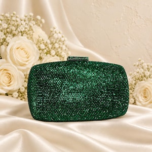 Puede incluir: Un bolso de mano rectangular completamente cubierto de peque&ntilde;os cristales de color verde esmeralda oscuro. El bolso cuenta con un cierre rectangular a juego, tambi&eacute;n incrustado con cristales verdes en la parte superior central.