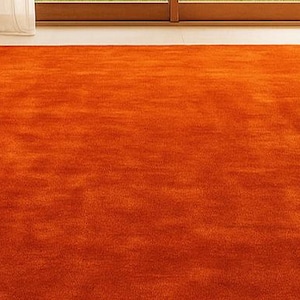 Op de afbeelding: Een groot, rechthoekig oranje tapijt met een getextureerd oppervlak. Het tapijt ligt op een lichtgekleurde vloer, met een bed en een raam zichtbaar op de achtergrond. De kleur van het tapijt is een levendige, warme tint.