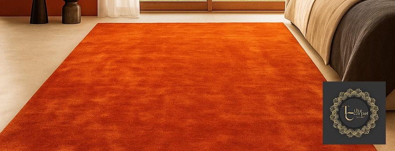 Op de afbeelding: Een groot, rechthoekig oranje tapijt met een getextureerd oppervlak. Het tapijt ligt op een lichtgekleurde vloer, met een bed en een raam zichtbaar op de achtergrond. De kleur van het tapijt is een levendige, warme tint.