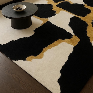 May include: A modern area rug with a bold abstract design. The rug features large black and gold shapes against a cream-colored background. The rug is placed on a dark wood floor, with a black coffee table and a cream-colored sofa in the background.