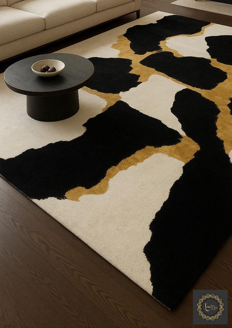 May include: A modern area rug with a bold abstract design. The rug features large black and gold shapes against a cream-colored background. The rug is placed on a dark wood floor, with a black coffee table and a cream-colored sofa in the background.