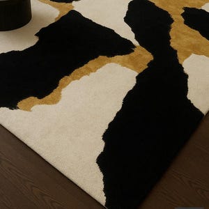 May include: A modern area rug with a bold abstract design. The rug features large, irregular shapes in black, cream, and gold. The rug's pattern creates a striking visual contrast, suitable for living rooms or bedrooms.