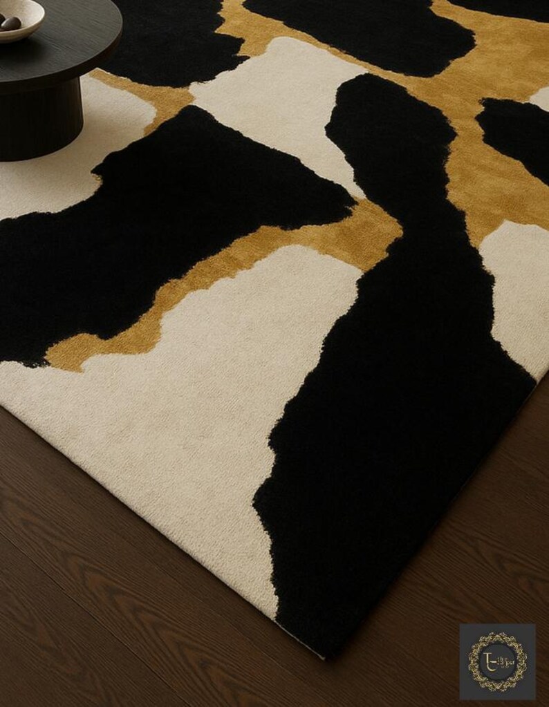 May include: A modern area rug with a bold abstract design. The rug features large, irregular shapes in black, cream, and gold. The rug's pattern creates a striking visual contrast, suitable for living rooms or bedrooms.