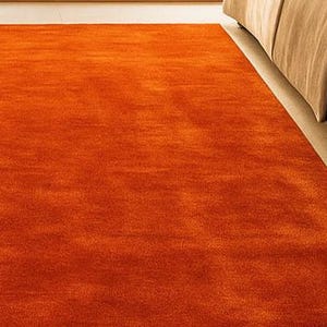 Op de afbeelding: Een feloranje vloerkleed met een zacht, getextureerd oppervlak. Het vloerkleed staat naast een bed met een bruin dekbed en een beige bedrok. De kleur van het vloerkleed voegt warmte toe aan de kamer.